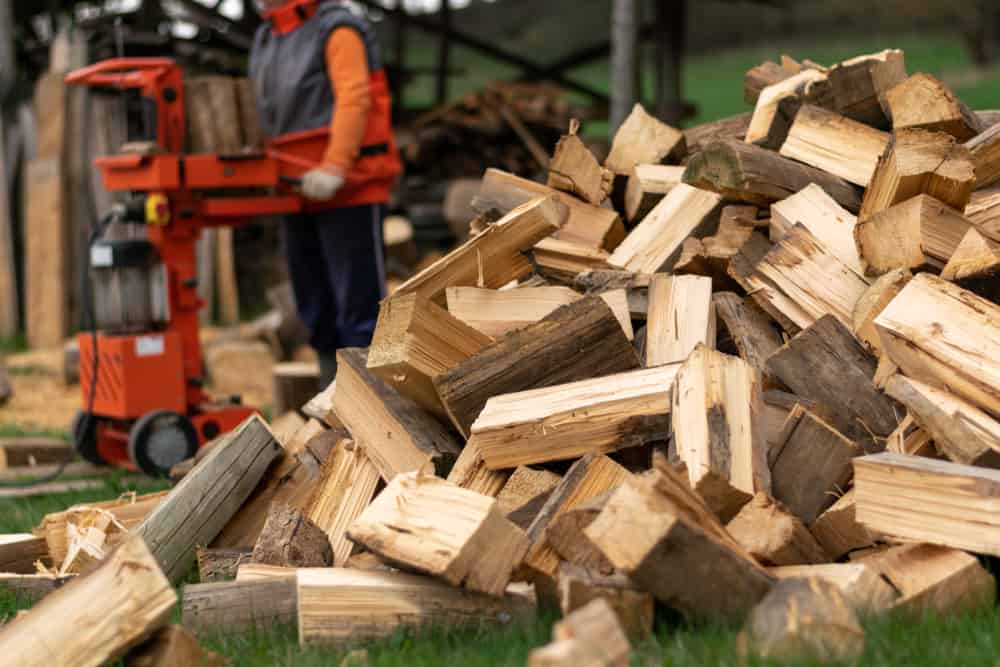 Pile of split wood with a man operating a wood splitter in the background illustrating understanding the wood splitter.