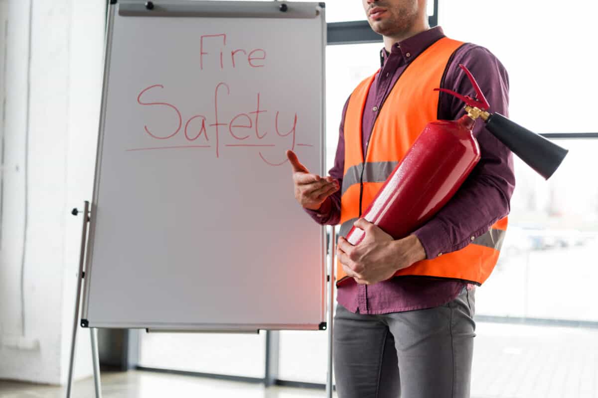 Cropped view of fireman holding red extinguisher while standing near white board with fire safety lettering