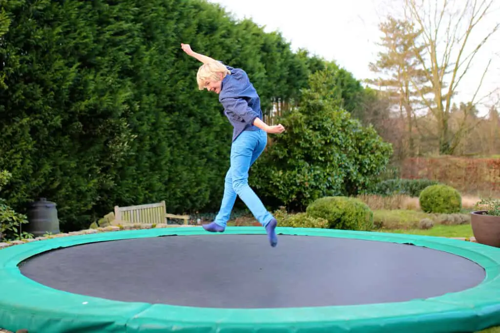 Happy kid plays outdoors in garden jumping high in the sky on trampoline. Active teenager boy having fun outdoors at early spring day.