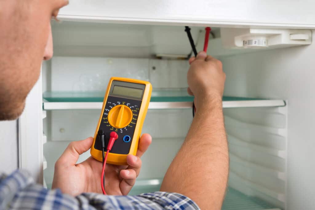 A Male Technician Checking Fridge With Digital Multimeter testing the differences between indoor and outdoor refrigerators.