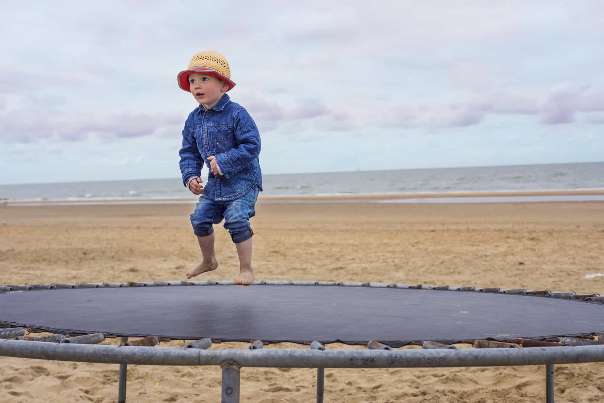 A child apprximately 2-year old jumping on a trampoline on a beach with water in background